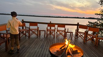 Ein Junge steht am Ufer eines ruhigen Gewässers, während ein Lagerfeuer in der Nähe flackert und der Sonnenuntergang den Himmel färbt.