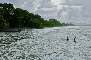 Zwei Männer stehen im flachen Wasser eines ruhigen Strandes, umgeben von üppigem Grün und sanften Wellen.