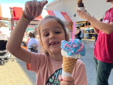 Ein fröhliches Kind hält ein buntes Eis in der Waffel und zeigt mit dem Finger nach oben, während es lächelt.