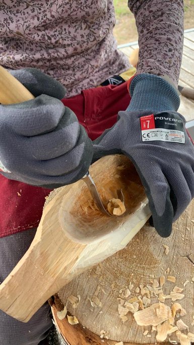 Eine Person schnitzt mit einem Werkzeug in ein Stück Holz, während Holzspäne um sie herum liegen.