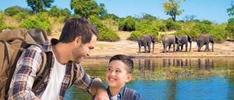 Ein Vater und sein Sohn lächeln sich am Ufer eines Gewässers an, während Elefanten im Hintergrund grasen.