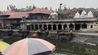 Zwei Männer in traditioneller Kleidung sitzen am Ufer des Bagmati-Flusses, umgeben von religiösen Gegenständen und Blumen.