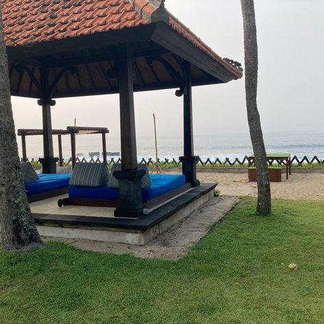 Ein traditioneller Pavillon mit blauen Liegen steht am Strand, umgeben von grünem Gras und Blick auf das ruhige Meer.