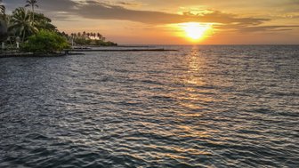 Ein atemberaubender Sonnenuntergang über dem ruhigen Wasser, umgeben von Palmen und einer friedlichen Küstenlandschaft.