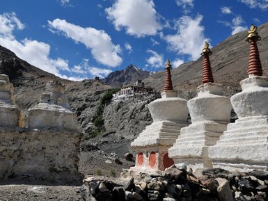 Drei weiße Stupas mit roten Spitzen stehen vor einer dramatischen Berglandschaft und einem Kloster im Hintergrund.