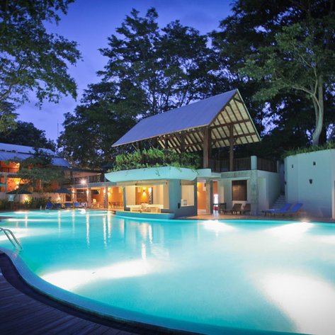 Ein ruhiger Poolbereich mit sanftem Licht, umgeben von Bäumen und einem modernen Gebäude im Hintergrund.