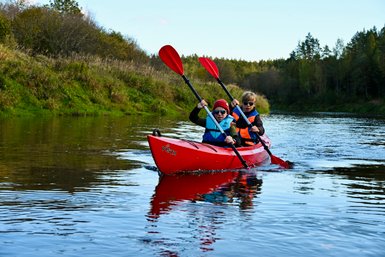 Zwei Kinder paddeln in einem roten Kajak auf einem ruhigen Fluss, umgeben von grünen Ufern und Bäumen im Hintergrund.
