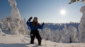 Eine fröhliche Frau in blauer Winterkleidung posiert im verschneiten Wald, umgeben von schneebedeckten Bäumen und strahlender Sonne.