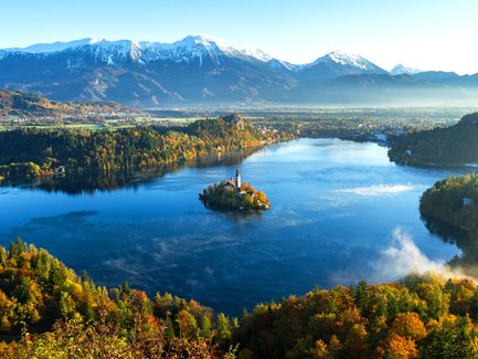 Ein malerischer Blick auf den Bleder See, umgeben von buntem Herbstlaub und majestätischen Bergen im Hintergrund.