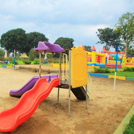 Ein bunter Spielplatz mit einer Rutsche in Rot und Lila, umgeben von grünen Bäumen und einem aufblasbaren Spielgerät im Hintergrund.