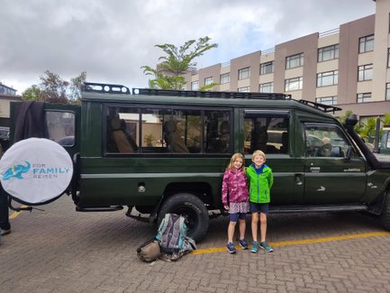 Zwei Kinder stehen lächelnd neben einem grünen Jeep, bereit für ein Abenteuer in Nairobi, mit einem Hotel im Hintergrund.