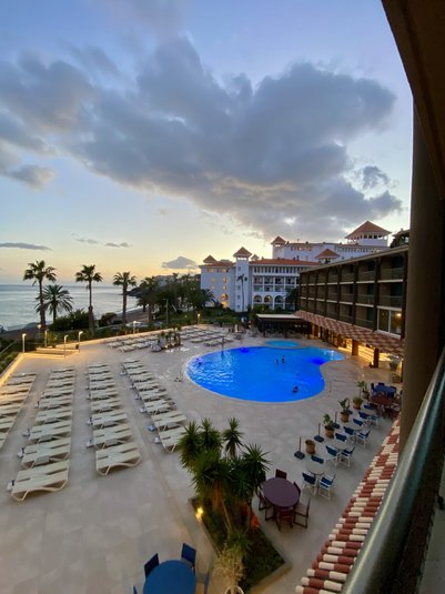 Ein luxuriöser Hotelpool mit Sonnenliegen, umgeben von Palmen und einem atemberaubenden Sonnenuntergang am Horizont.