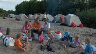 Eine Gruppe von Kindern und Erwachsenen sitzt um ein Lagerfeuer am Strand, umgeben von Zelten und Bäumen im Hintergrund.