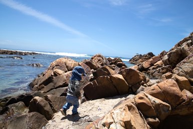 Entdeckungsfreudiges Baby geht vorsichtig über die Felsen an der Küste von Mossel Bay – Garden Route mit Kindern
