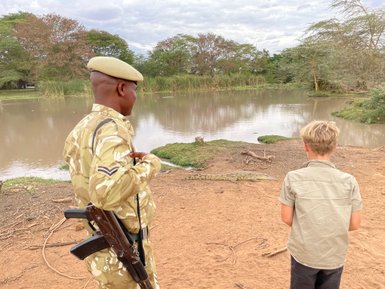Ein Sicherheitsbeamter in Uniform beobachtet einen Jungen, der fasziniert auf einen Krokodil am Ufer schaut.