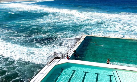 Der Pool am Bondi Beach reflektiert das Sonnenlicht, während Wellen an den Rand schlagen und Menschen schwimmen.