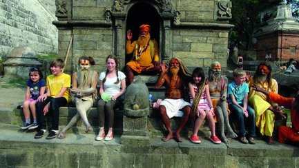 Eine Gruppe von Kindern und heiligen Männern sitzt auf einer Steinbank vor einem Tempel in Nepal, alle lächeln freundlich.