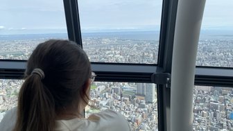 Eine Person mit einem Pferdeschwanz blickt aus dem Fenster des Tokio Skytree auf die weitläufige Stadtlandschaft.