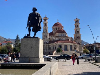 Eine Statue eines historischen Mannes steht vor einer beeindruckenden Kirche mit roten Kuppeln und einem klaren blauen Himmel.