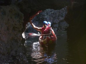 Zwei Personen in einem roten Schlauchboot paddeln durch eine dunkle Höhle, umgeben von glatten Felsen und Wasser.