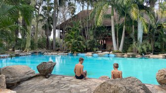Zwei Jungen sitzen am Rand eines blauen Pools, umgeben von üppigen Palmen und Felsen, während sie die Aussicht genießen.