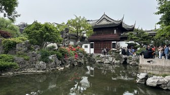 Ein traditionelles chinesisches Gartenhaus mit geschwungenem Dach, umgeben von üppigem Grün und bunten Blumen am Teich.