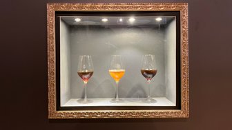 Drei elegante Weingläser stehen in einer beleuchteten Vitrine, gefüllt mit verschiedenen Getränken in warmen Farbtönen.