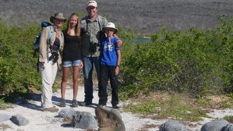 Eine Familie steht lächelnd auf einem sandigen Strand, während ein Seelöwe in der Nähe entspannt sitzt.