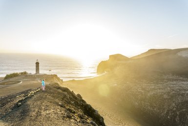 Eine Person steht auf einem Hügel und blickt auf den Ozean, während die Sonne hinter dem Leuchtturm aufgeht.