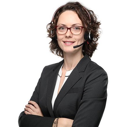 Eine lächelnde Frau mit lockigem Haar trägt ein Headset und einen eleganten Anzug, während sie selbstbewusst posiert.