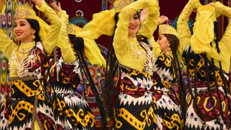 Fünf Tänzerinnen in bunten traditionellen Kleidern bewegen sich rhythmisch zur Musik, ihre Hände heben sich in die Luft.