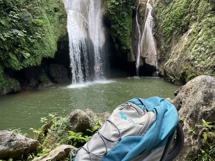Ein blauer Rucksack steht auf einem Felsen, während ein Wasserfall im Hintergrund in einen klaren Teich fließt.