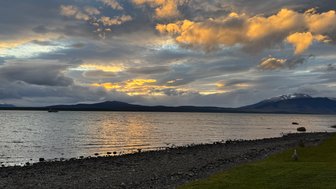 Ein malerischer Sonnenuntergang über dem Wasser, mit dramatischen Wolken und sanften Wellen, die den Uferbereich umspielen.