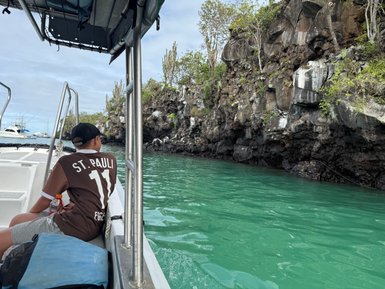 Ein Junge sitzt auf einem Boot und blickt auf die grüne Wasseroberfläche und die felsige Küste im Hintergrund.