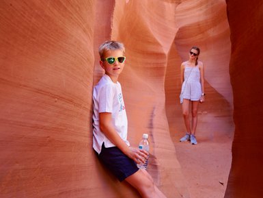Ein Junge mit Sonnenbrille lehnt an einer roten Felswand und hält eine Wasserflasche, während ein Mädchen im Hintergrund steht.