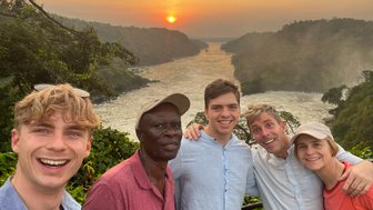 Fünf Personen posieren fröhlich vor einer atemberaubenden Landschaft mit einem Sonnenuntergang über einem Fluss.