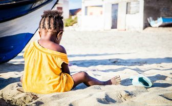 Ein kleines Kind sitzt im Sand, schaut auf einen Strand, mit einem Boot im Hintergrund und einem leuchtend gelben T-Shirt.