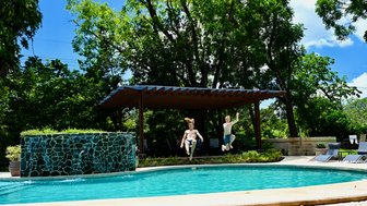 Zwei Jungen springen gleichzeitig ins erfrischende Wasser eines Pools, umgeben von üppigem Grün und einer Sonnenterrasse.