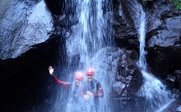 Zwei Personen in Neoprenanzügen stehen unter einem Wasserfall und lächeln, während Wasser über sie herabfließt.
