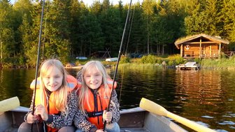 Zwei fröhliche Mädchen in orangefarbenen Schwimmwesten sitzen im Boot und halten Angelruten in der Hand.