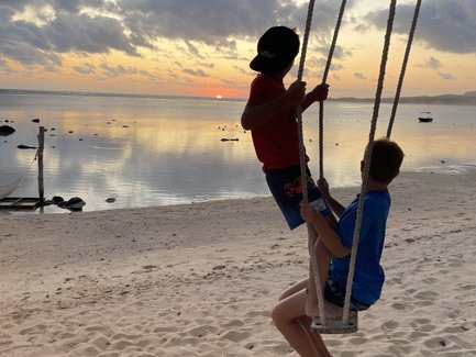 Zwei Kinder schwingen fröhlich auf einer Schaukel am Strand, während die Sonne hinter dem Horizont untergeht.