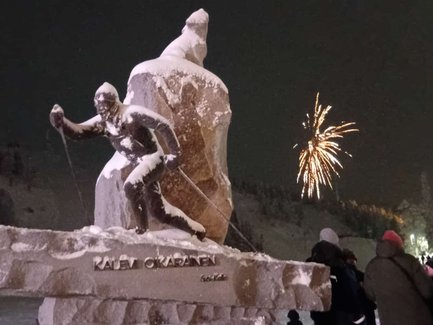 Eine schneebedeckte Statue eines Skifahrers steht im Vordergrund, während Feuerwerk den Nachthimmel erleuchtet.