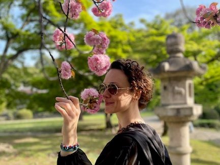 Eine Frau mit Sonnenbrille steht unter blühenden Kirschbäumen und hält einen Zweig mit rosa Blüten in der Hand.