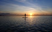 Eine Person paddelt auf einem Stand-Up-Paddle-Board bei Sonnenuntergang, während die Wellen sanft glitzern.