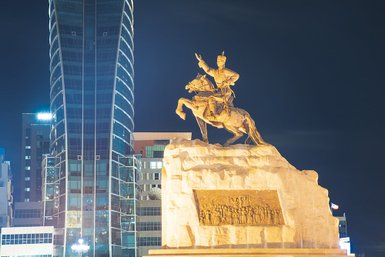 Eine goldene Statue eines Reiters, der auf einem Felsen steht, umgeben von modernen Gebäuden bei Nacht.