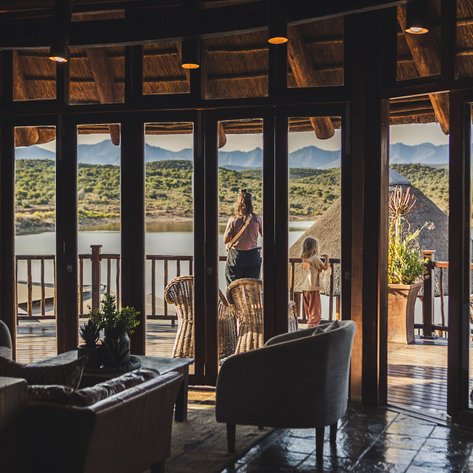 Eine Frau und ein Kind stehen am Fenster und genießen die Aussicht auf den ruhigen See und die umliegenden Berge.