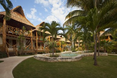 Ein tropischer Garten mit Palmen und einem kleinen Teich, umgeben von rustikalen Hütten mit Strohdächern.