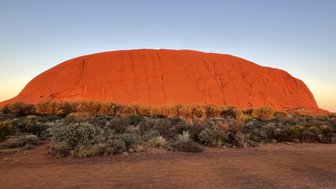 Der majestätische Ayers Rock leuchtet in warmen Rottönen während der Abenddämmerung, umgeben von üppiger, wilder Vegetation.