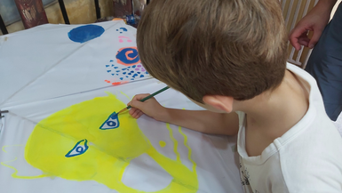 Ein Kind malt mit einem Pinsel auf einem großen weißen Blatt, das eine leuchtend gelbe Kreatur darstellt.
