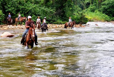 Eine Gruppe von Reitern durchquert einen klaren Fluss, umgeben von üppigem, grünem Dschungel und Felsen.